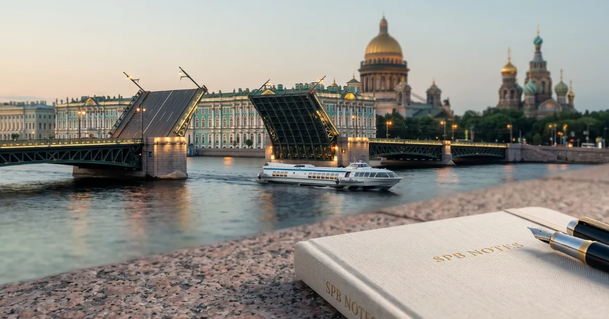 Крупный план светлого холщового блокнота с тиснением «SPB NOTES» и двух перьевых ручек, лежащих на гранитной набережной. На заднем плане, через реку Неву в сумерках, виден культовый силуэт Санкт-Петербурга: разведенный Дворцовый мост, бирюзовый Зимний дворец (Эрмитаж), золотой купол Исаакиевского собора и разноцветные купола Спаса на Крови. По воде проходит речной трамвайчик.